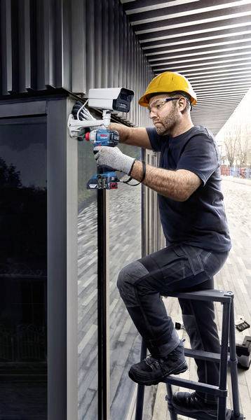 Ein Arbeiter auf einer Leiter installiert eine Überwachungskamera an einem Gebäude. Er trägt einen gelben Schutzhelm und Arbeitshandschuhe.