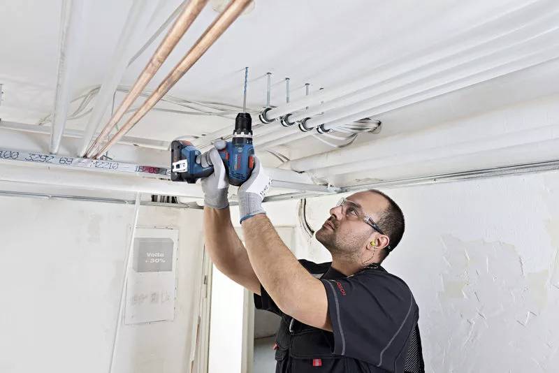 Ein Arbeiter, der mit einer Bohrmaschine mehrere Kabel und Rohre an einer Decke befestigt. Er trägt Schutzkleidung und Handschuhe.