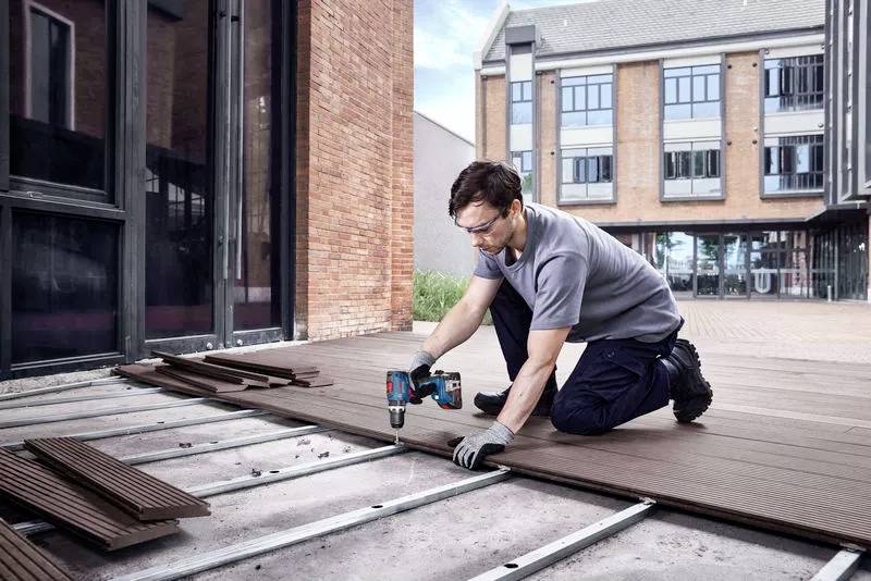 Ein Mann kniet auf einer Terrasse und verlegt Holzdielen mithilfe einer Bohrmaschine. Im Hintergrund sind moderne Gebäude zu sehen.
