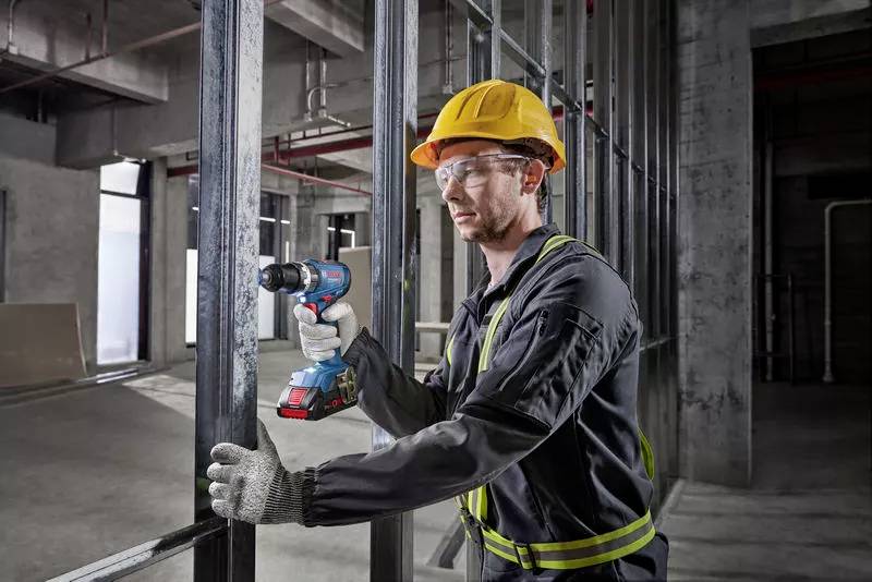 Ein Bauarbeiter mit Schutzhelm und Handschuhen benutzt eine Bohrmaschine, um Metallrahmen in einem Rohbau zu befestigen.