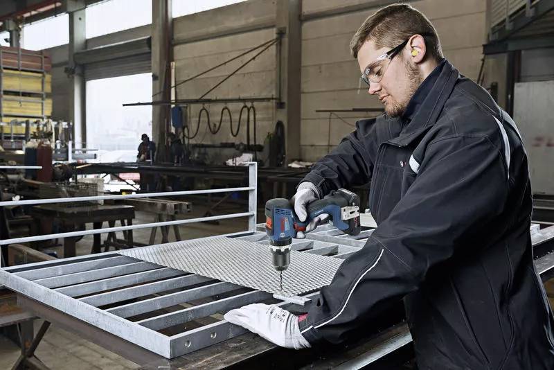 Ein Mann in einer Werkstatt bohrt mit einem Elektrowerkzeug ein Loch in ein Metallgitter, umgeben von industriellen Geräten.
