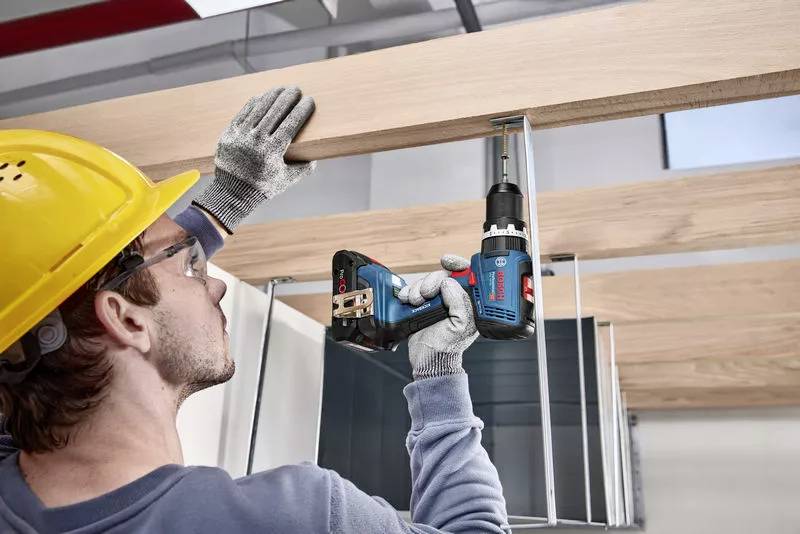 Arbeiter mit gelbem Schutzhelm und Handschuhen befestigt Holzleiste mit einer blauen Bohrmaschine an einer Deckenstruktur.