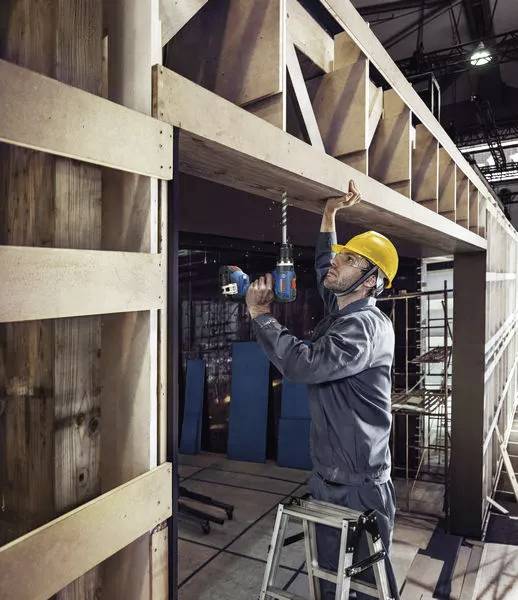 Ein Bauarbeiter mit gelbem Schutzhelm steht auf einer Leiter und schraubt mit einer Bohrmaschine an einer Holzkonstruktion in einer Halle.