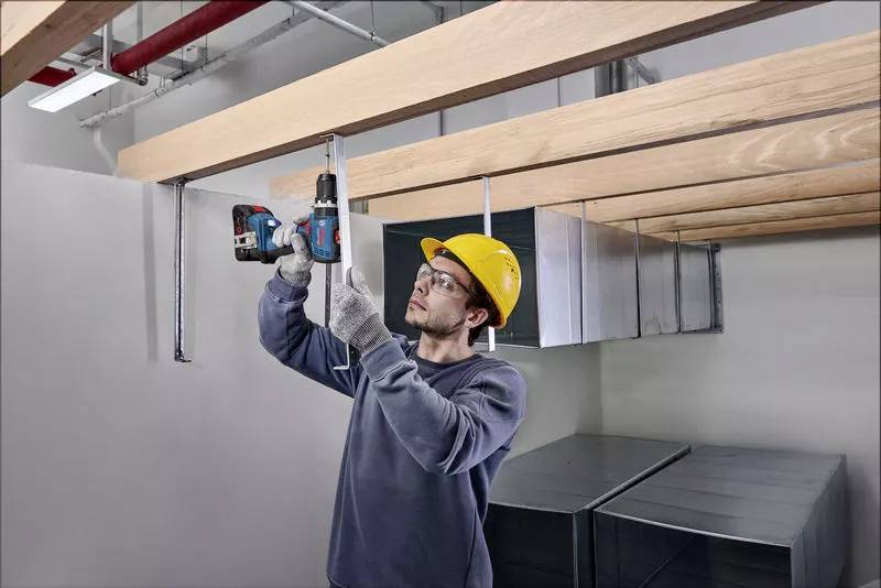 Ein Arbeiter mit Schutzhelm befestigt Holzbalken an der Decke mit einer Bohrmaschine in einem industriellen Innenraum.