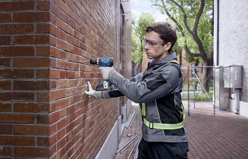 Ein Arbeiter bohrt mit einer elektrischen Bohrmaschine in eine Ziegelwand, trägt Sicherheitsbrille und Schutzausrüstung.
