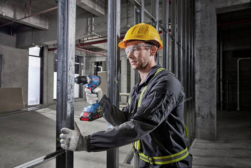 Ein Bauarbeiter mit Schutzhelm und Handschuhen bohrt mit einer Elektroschraubmaschine in Metallstützen auf einer Baustelle.