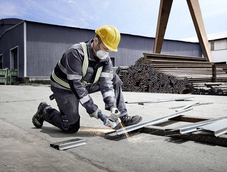 Ein Bauarbeiter mit Helm und Schutzmaske schneidet Metall mit einem Winkelschleifer auf einer Baustelle. Im Hintergrund sind Stapel von Metallbalken sichtbar.