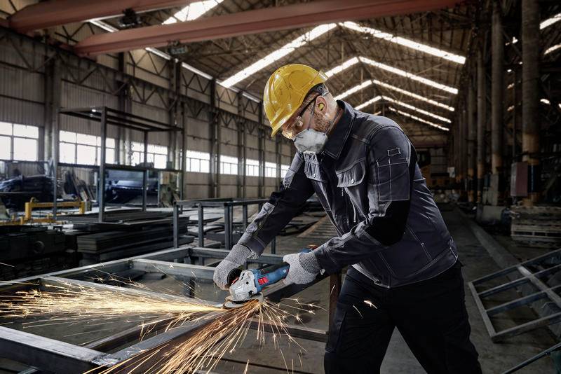Ein Arbeiter in Schutzkleidung schleift Metall mit einem Winkelschleifer in einer großen Fabrikhalle, Funken sprühen.