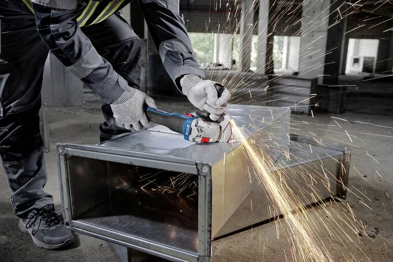 Eine Person schneidet mit einer Winkelschleifmaschine Metall. Funken fliegen, was auf intensive Arbeit hindeutet. Im Hintergrund Baustellenumgebung.