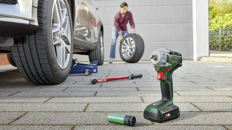 Eine Person wechselt ein Autoreifen in einer Einfahrt. Im Vordergrund liegt ein grüner Akkuschrauber auf dem Boden.