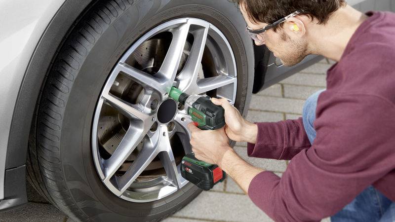 Ein Mann in Schutzbrille zieht mit einem Akkuschrauber eine Radmutter an einem Autoreifen fest. Der Reifen ist auf grauen Steinen.