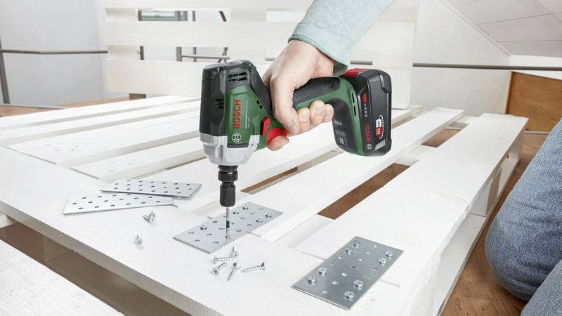 Eine Person schraubt Metallplatten auf eine weiße Holzpalette mit einem Bosch-Akkuschrauber fest. Auf der Palette liegen Schrauben.