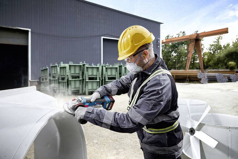 Eine Person mit Schutzhelm und Maske schleift ein großes Metallteil in einem Industriegebiet. Im Hintergrund sind Lagerhallen zu sehen.