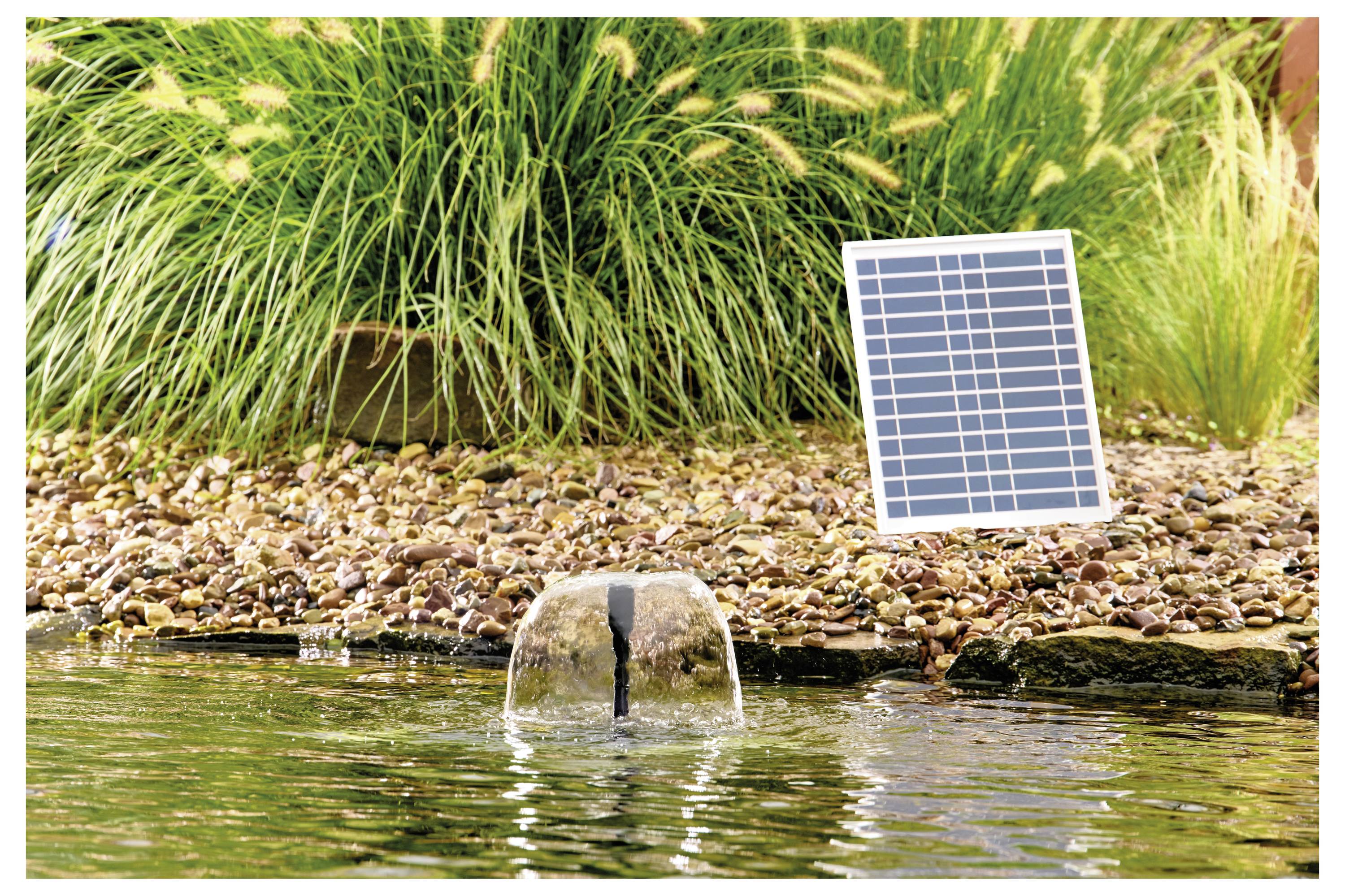 Ein kleines Solarpanel betreibt einen Brunnen in einem Teich, umgeben von Steinen und hohen Gräsern. Der Brunnen erzeugt einen sanften Wassersprühnebel.