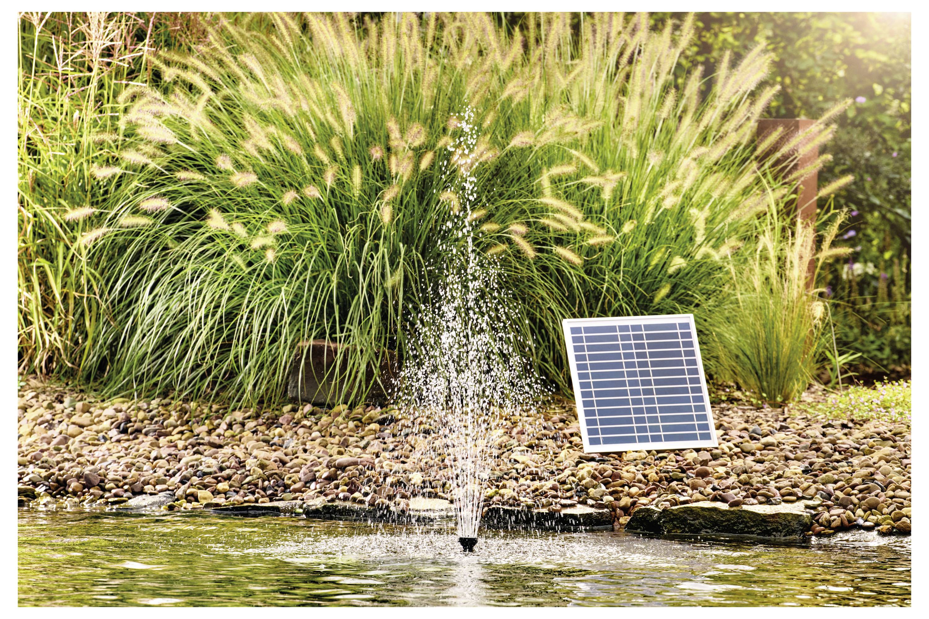 Ein kleiner Teichbrunnen, umgeben von Steinen, der durch ein Solarpanel betrieben wird, das neben ihm auf dem Boden platziert ist, mit hohem Gras im Hintergrund.