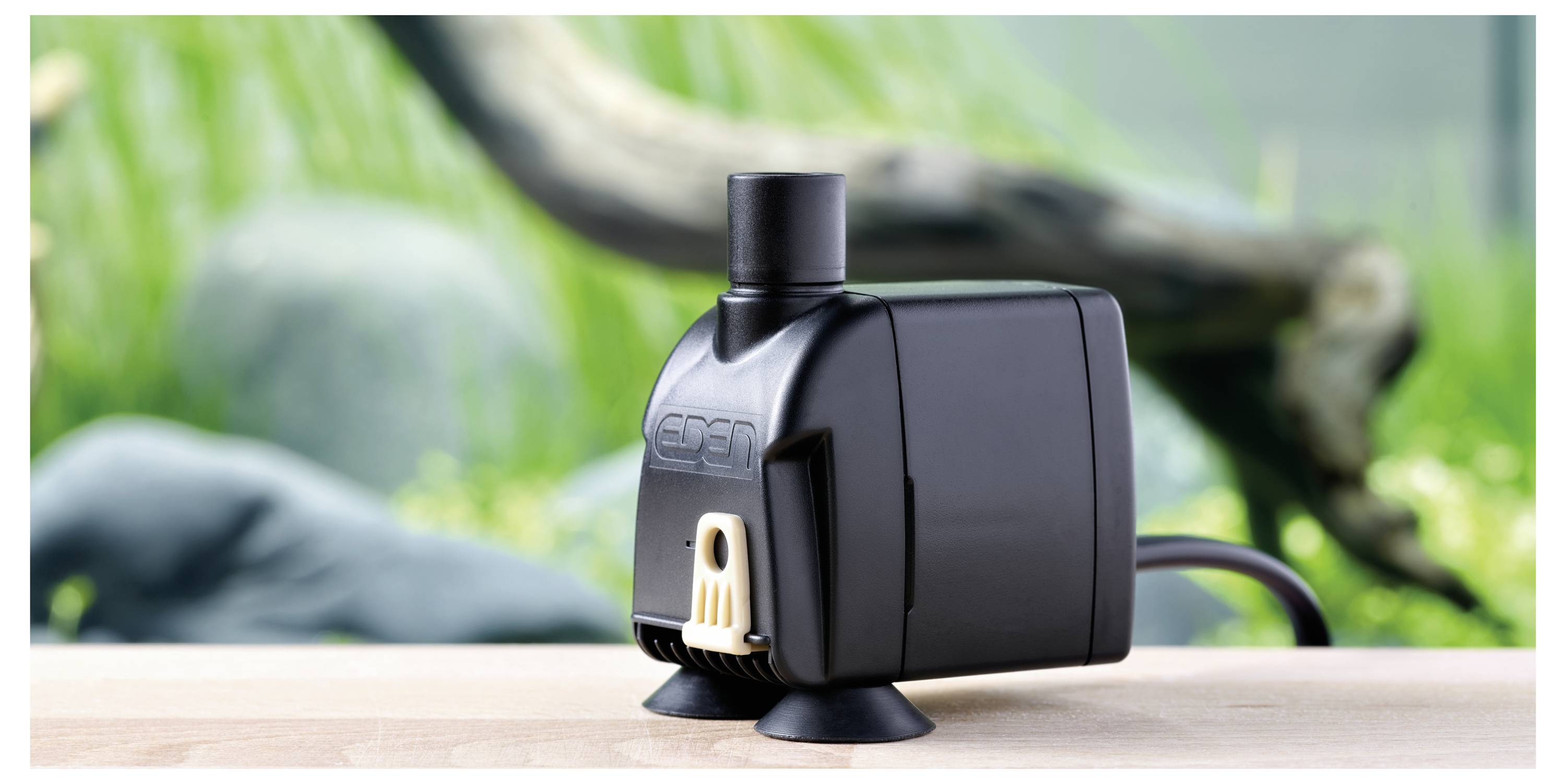 Schwarze Aquarium-Wasserpumpe auf einer Holzoberfläche, mit im Hintergrund unscharf verschwommenen grünen Wasserpflanzen und Treibholz.