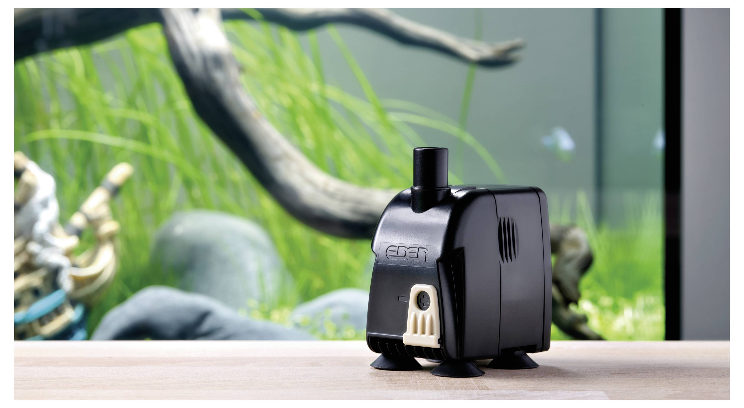 Eine kleine schwarze Aquarienpumpe steht auf einer Holzoberfläche vor einem Aquariumhintergrund mit grünen Pflanzen und Treibholz.