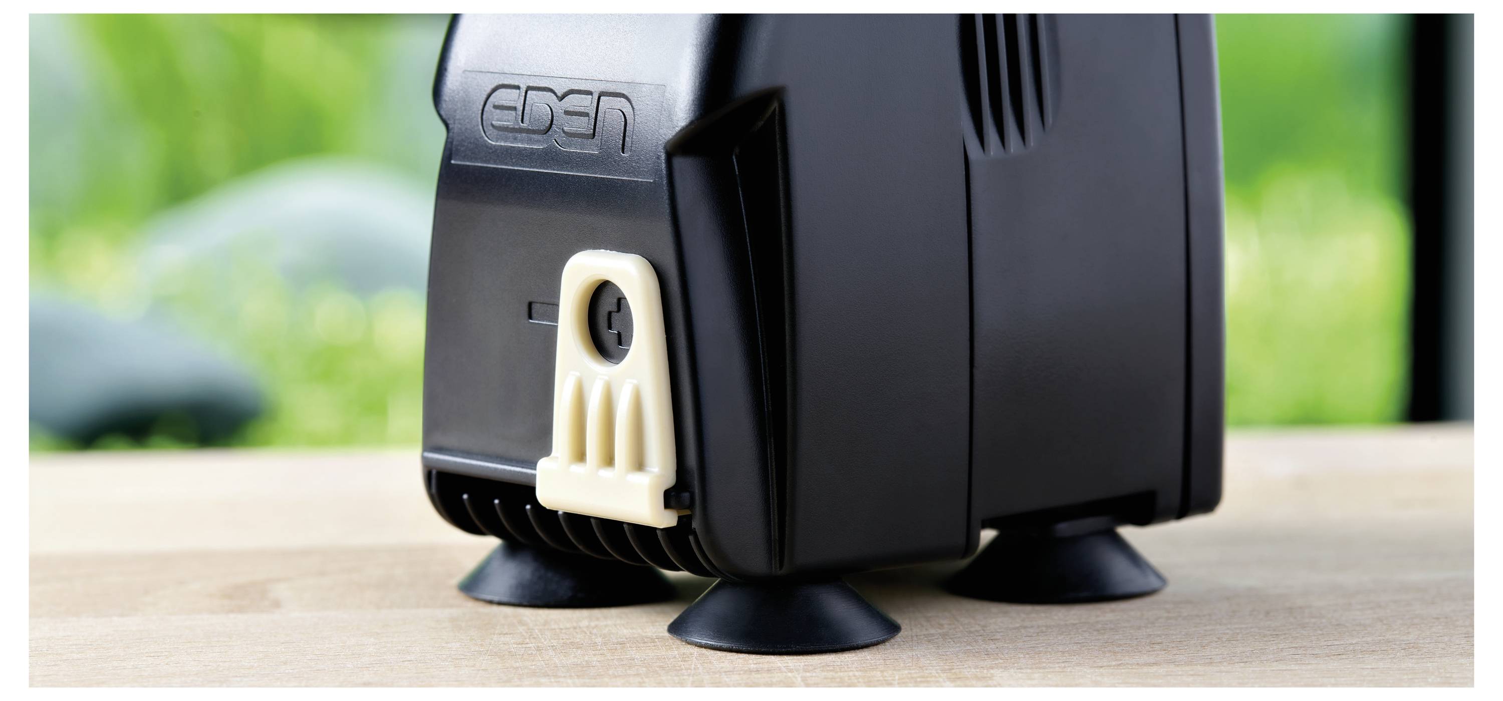 Ein schwarzes Gerät mit einem beigen Clip, das auf vier Saugnapffüßen ruht, ist auf einer Holzoberfläche vor einem unscharfen grünen Hintergrund platziert.