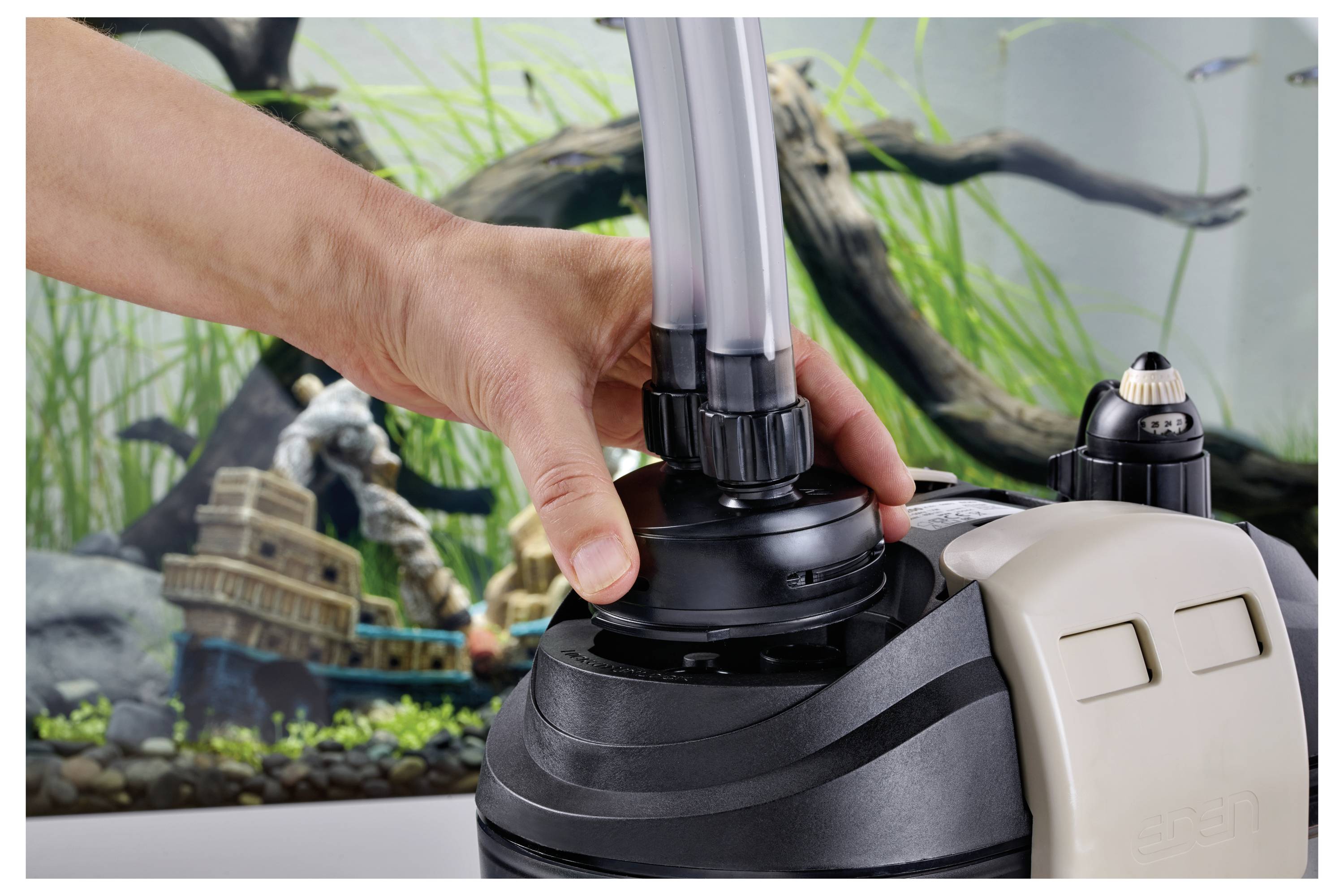 Eine Hand montiert eine Filtereinheit, die mit Schläuchen vor einem Aquarium mit Pflanzen und dekorativen Strukturen verbunden ist.