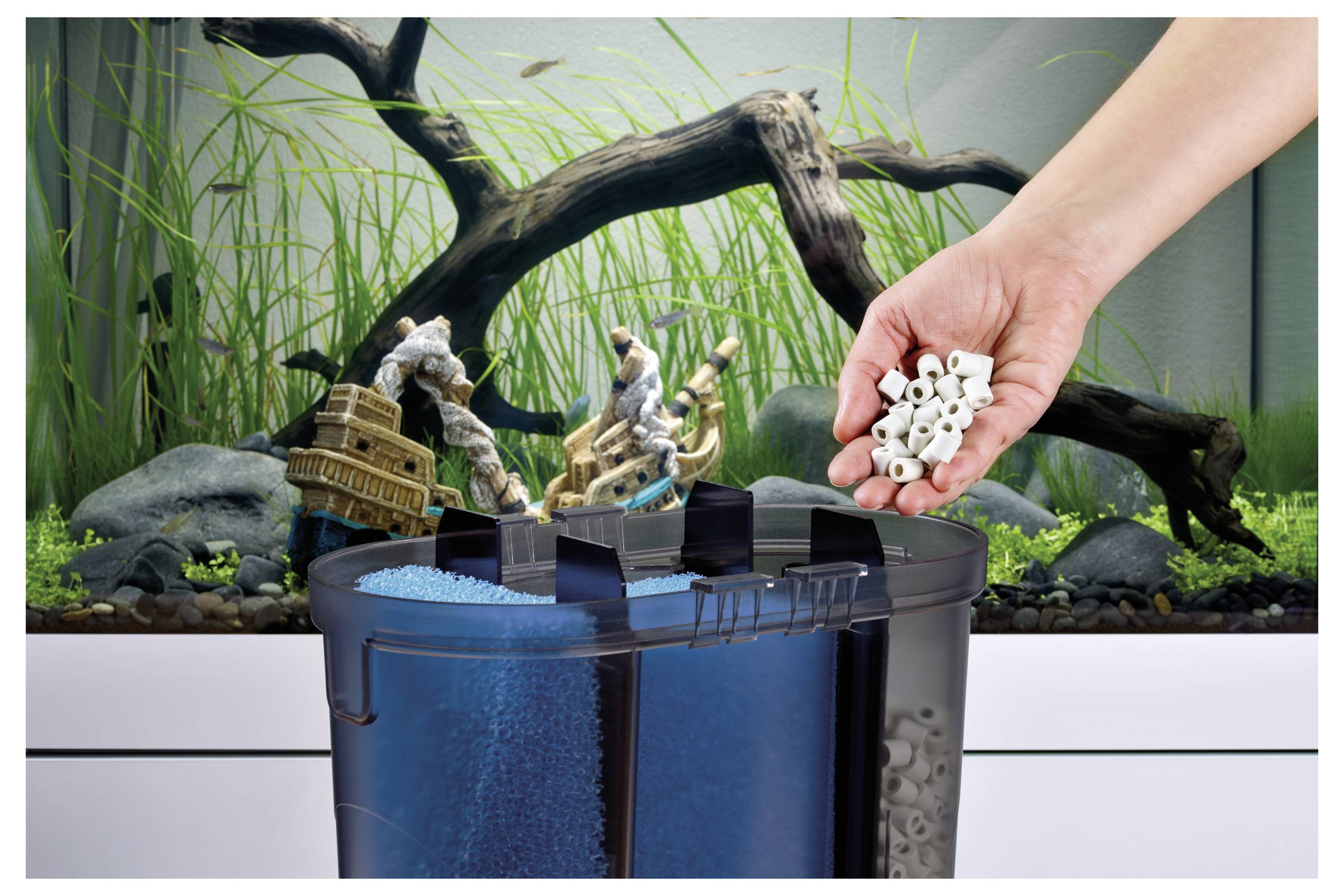 Eine Hand, die Filtermedien über einem Aquariumfilter hält, mit einem Fischbecken-Hintergrund aus Pflanzen und Dekorationen.