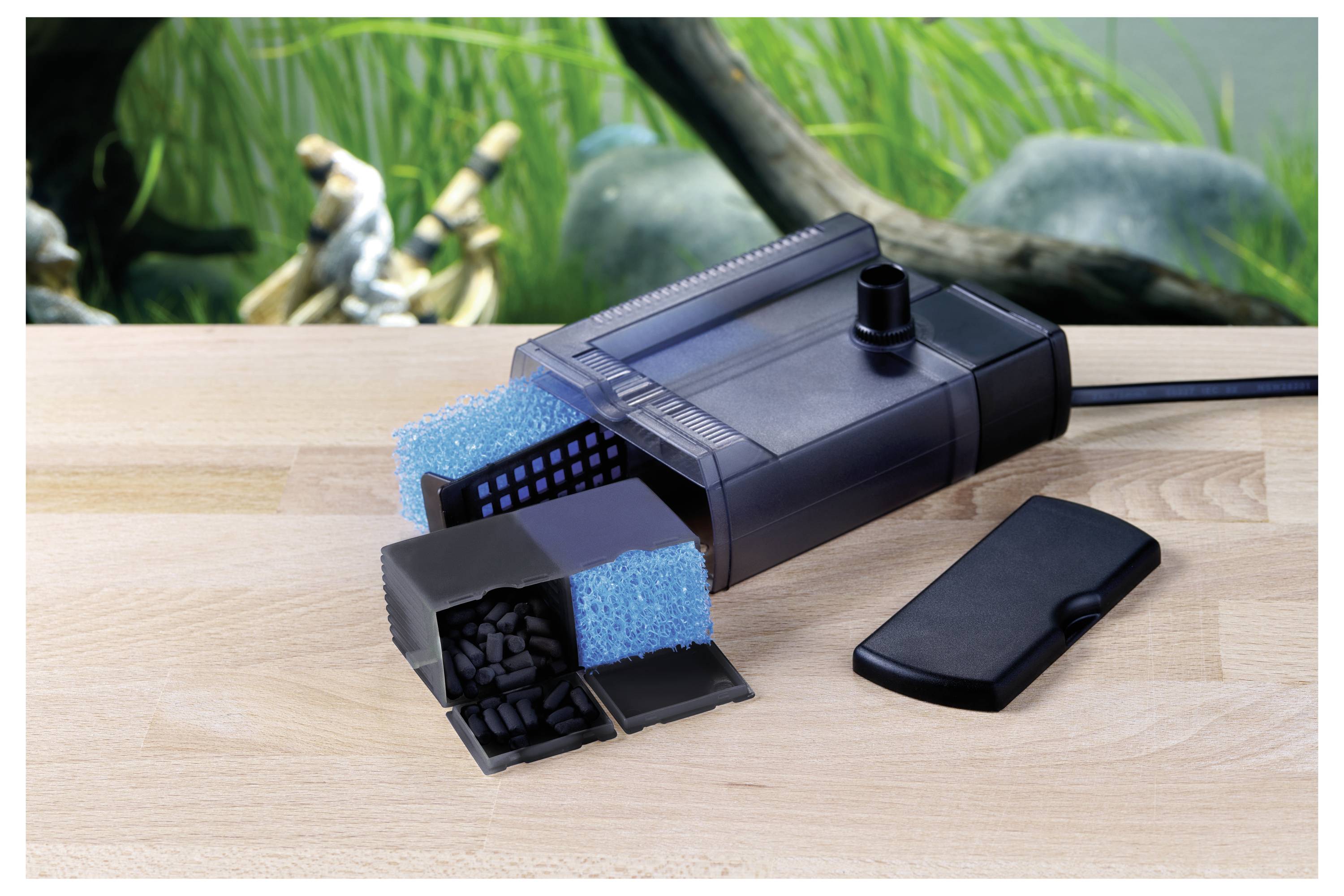 Ein Aquariumfilter mit Aktivkohle- und Schaumkomponenten wird zerlegt auf einer Holzoberfläche vor einem Aquarienhintergrund dargestellt.
