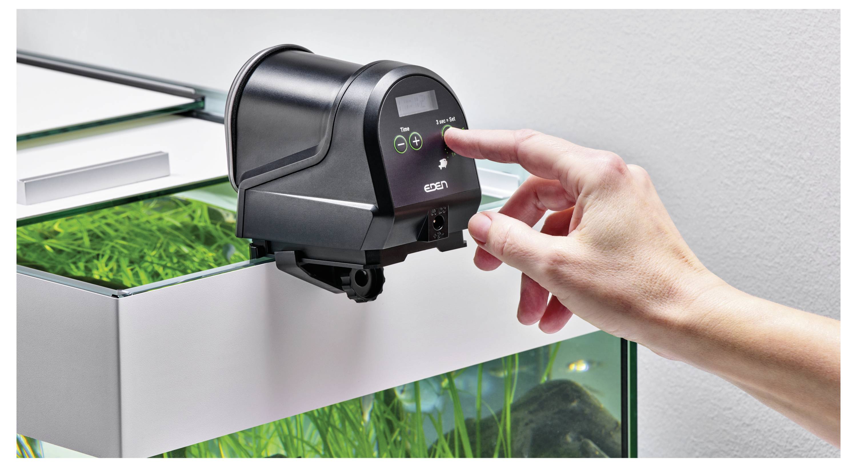 Eine Person stellt einen digitalen Timer an einem automatischen Fischfütterungsgerät ein, das am Rand eines Aquariums mit grünen Pflanzen befestigt ist.