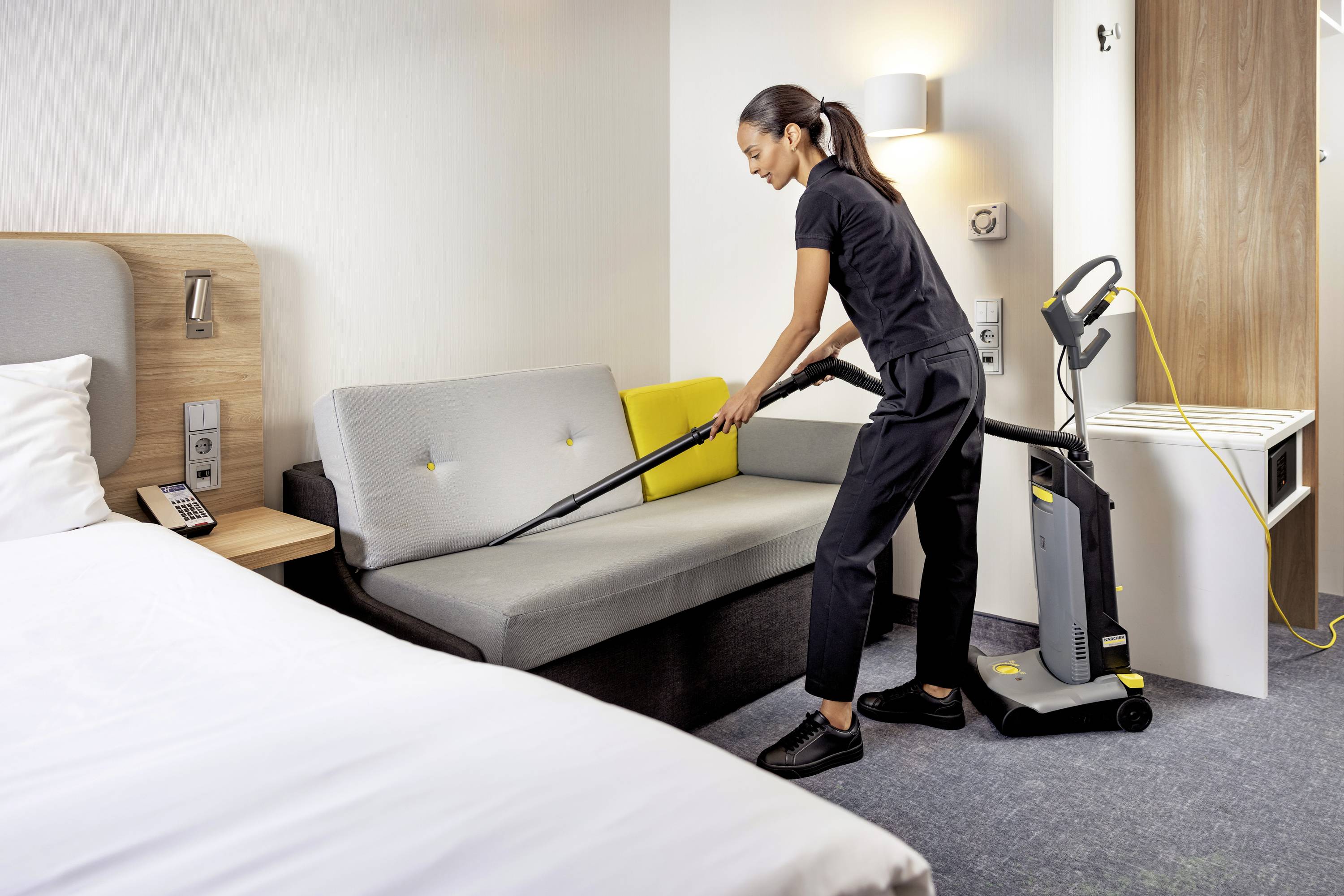 Eine Person reinigt ein Sofa in einem Hotelzimmer mit einem Staubsauger. Im Vordergrund ist ein Bett zu sehen.
