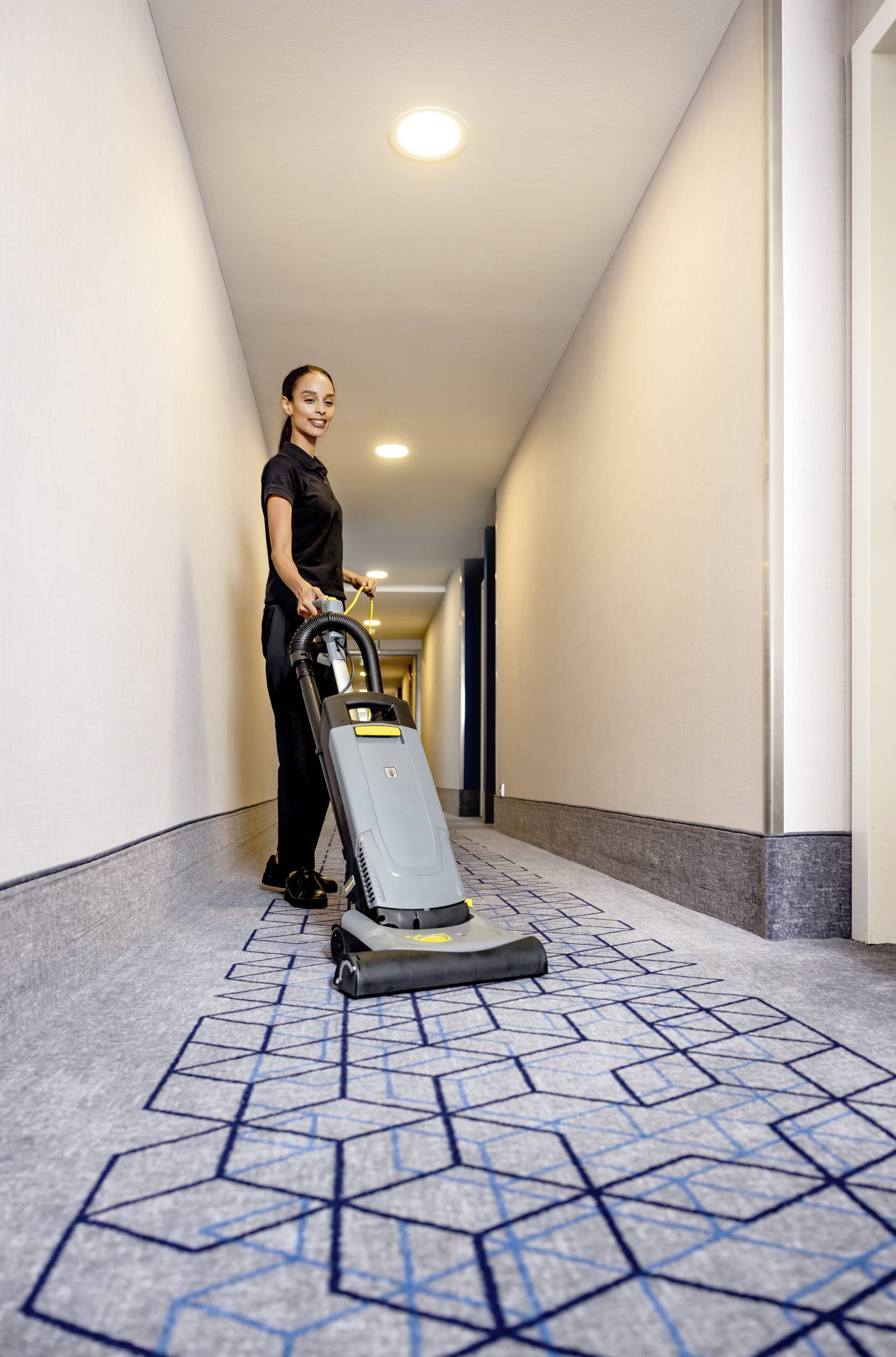 Eine Frau reinigt mit einem Staubsauger einen Flur mit geometrisch gemustertem Teppichboden.