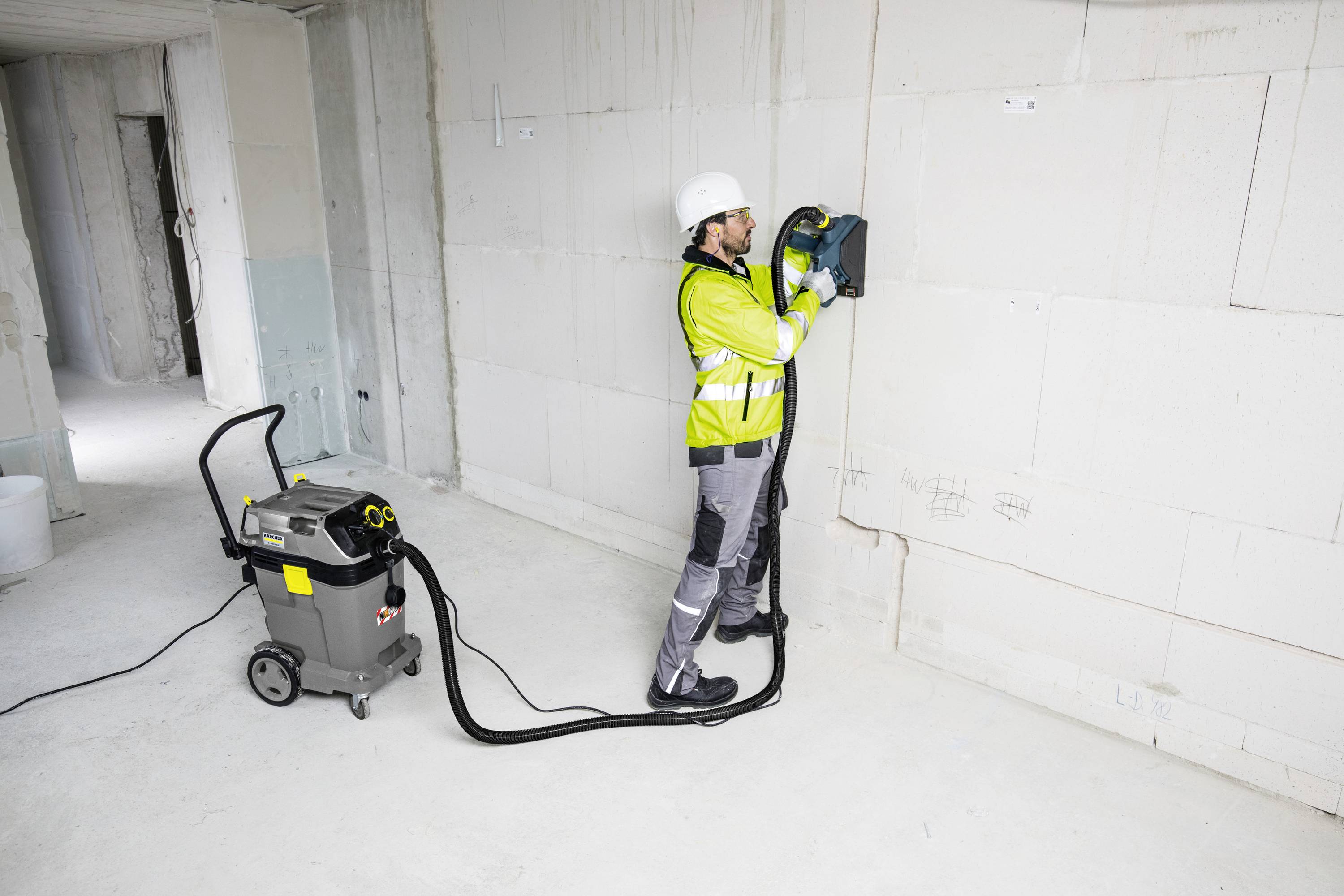 Ein Bauarbeiter in Schutzkleidung bedient eine Mauerfräse an einer Wand in einem unfertigen Raum. Ein Staubsauger ist angeschlossen.