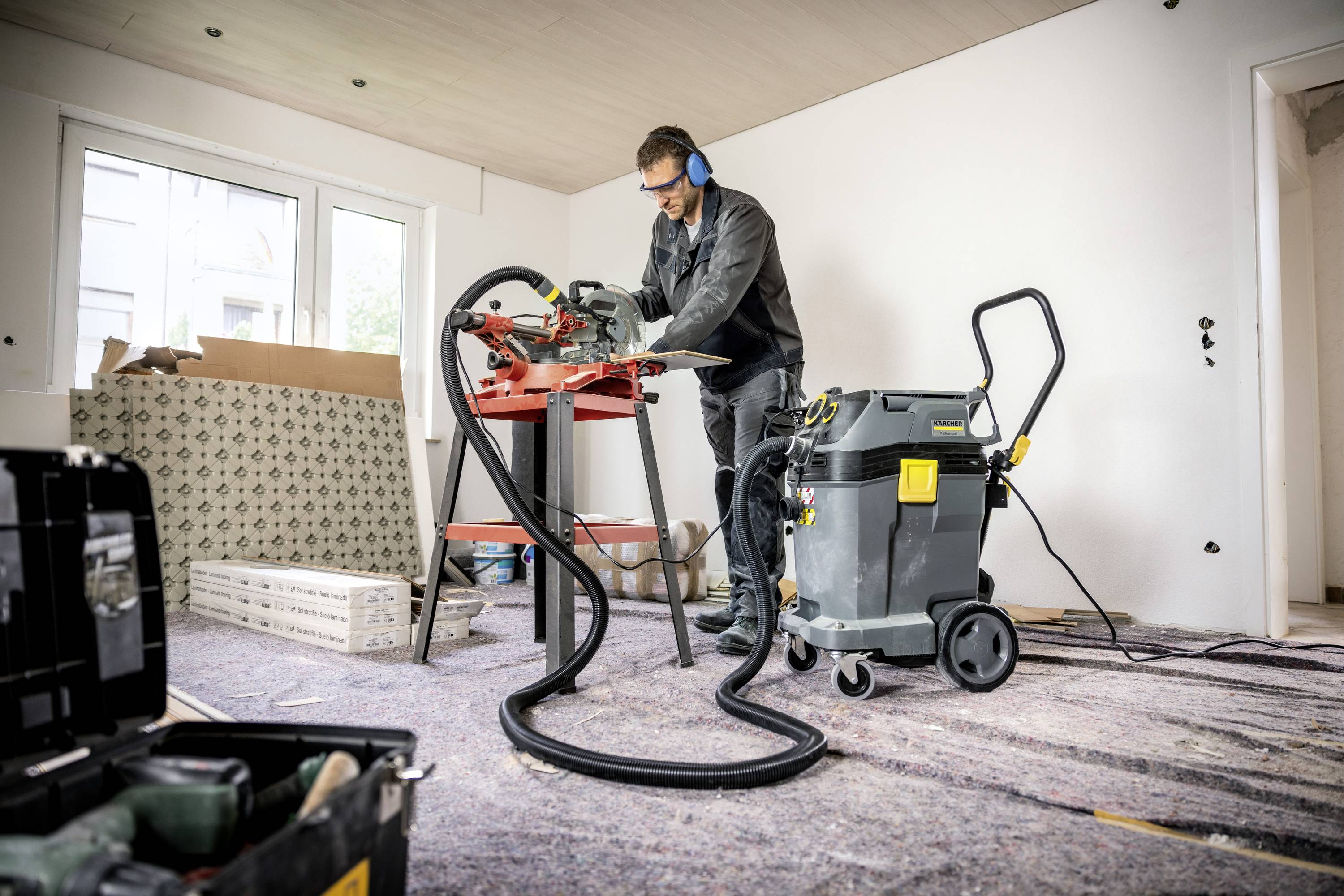 Ein Mann arbeitet in einem unfertigen Raum mit einer Säge auf einem Tisch. Er trägt Schutzkleidung und die Umgebung wirkt wie eine Baustelle.