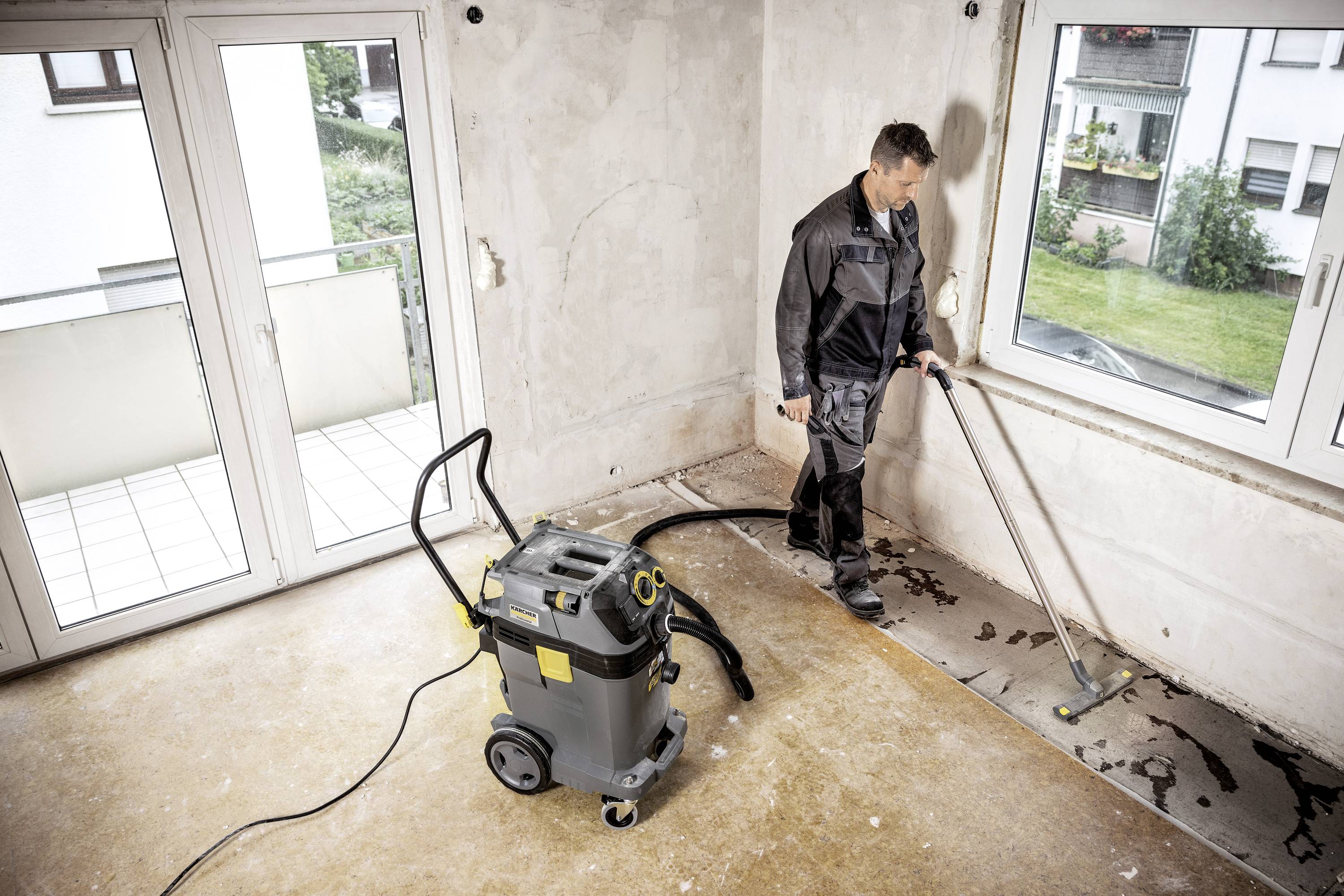 Ein Mann reinigt einen leeren Raum mit einem Wassersauger. Der Boden ist teilweise feucht und das Fenster zeigt einen Gartenblick.