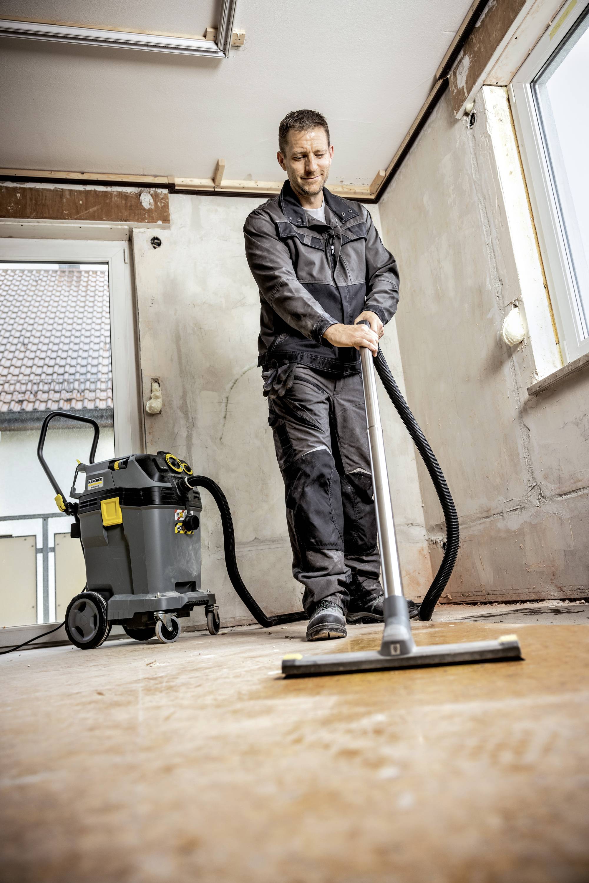 Eine Person in Arbeitskleidung reinigt den Boden eines leeren Raumes mit einem Industriesauger. Der Raum wirkt renovierungsbedürftig.