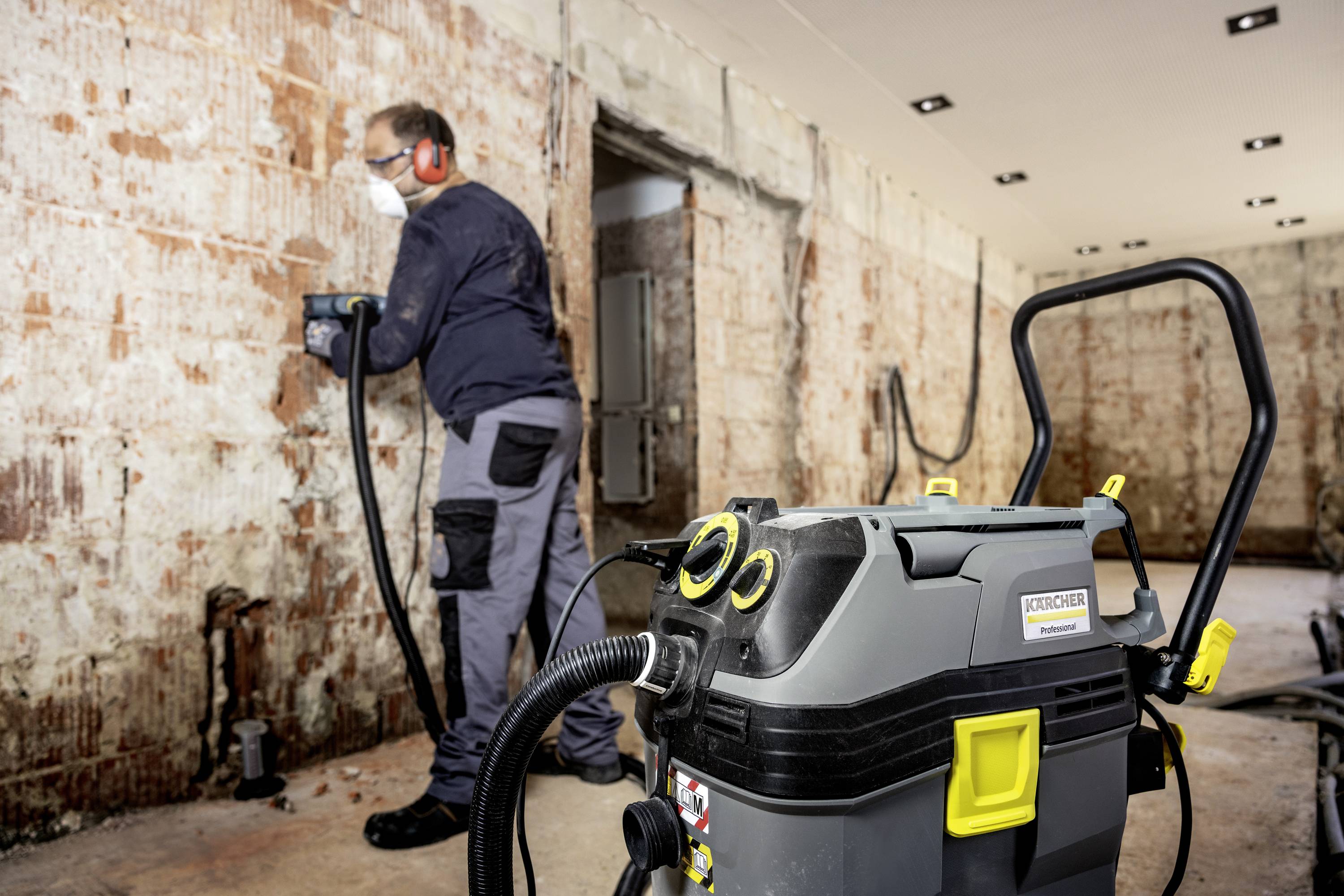 Ein Handwerker entfernt alten Putz von einer Wand mit einem Bohrhammer. Im Vordergrund steht ein Industriesauger, der Staub aufsaugt.