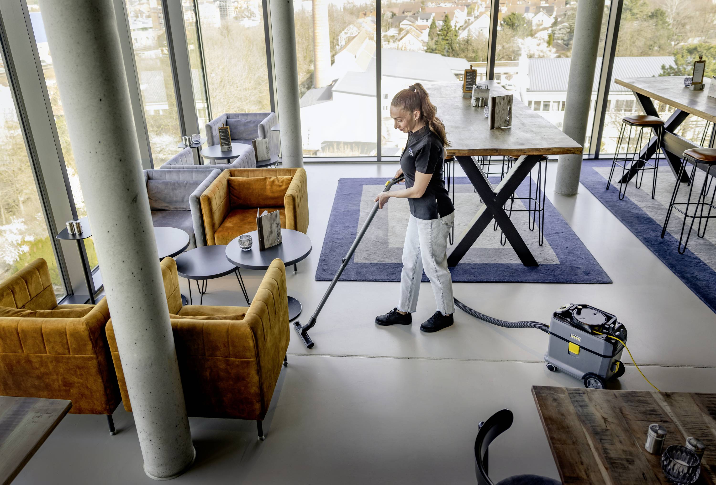 Eine Person staubsaugt den Boden in einem modernen, lichtdurchfluteten Café mit großen Fenstern, Holztischen und gemütlichen Sesseln.