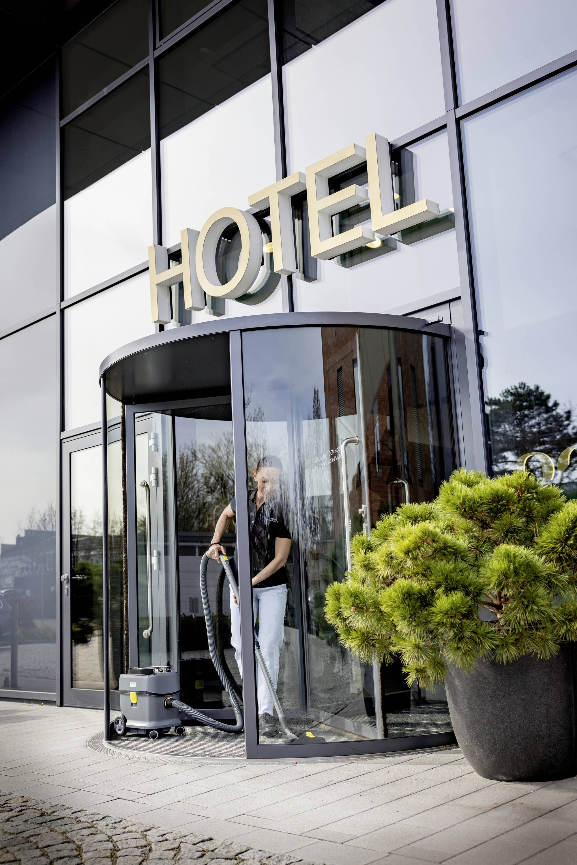 Eine Person reinigt den Eingang eines modernen Hotels mit Glastüren. Große goldene Buchstaben über der Tür formen das Wort 'HOTEL'.