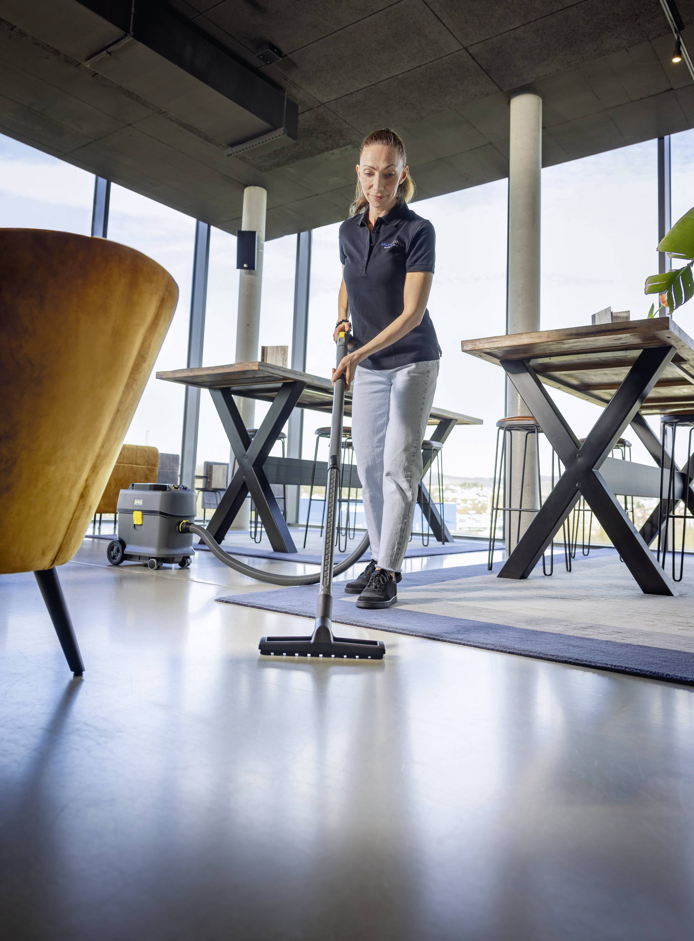 Eine Person reinigt den Boden eines modernen, hellen Raumes mit einem Staubsauger. Im Hintergrund sind Tische und große Fenster sichtbar.
