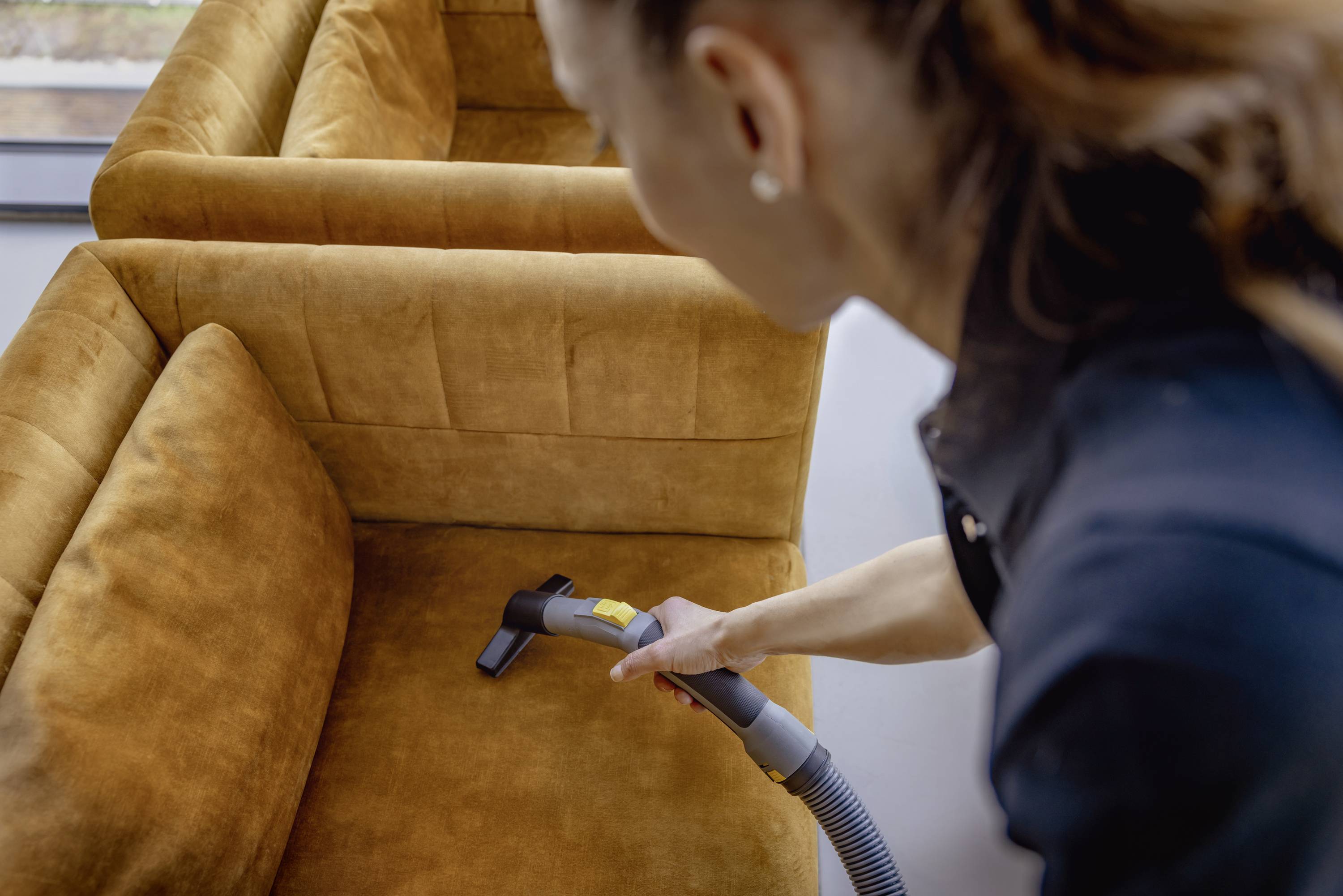 Eine Person reinigt ein goldenes Polstersofa mit einem Staubsauger, Fokus auf die Reinigungsdüse und den Stoff.