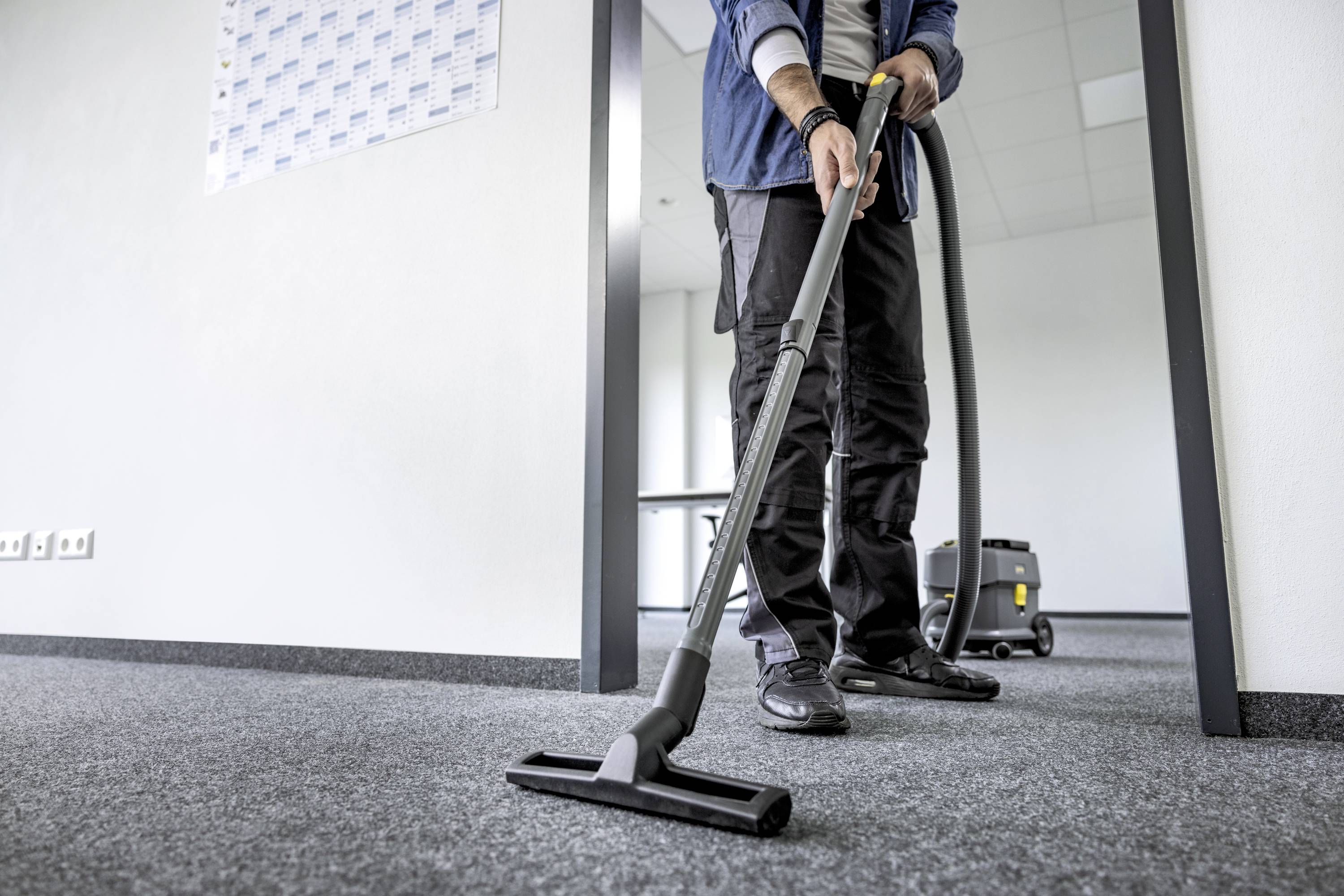 Person, die einen Staubsauger benutzt, um den Teppichboden in einem Büro zu reinigen. Im Hintergrund sind Möbel und eine Wand zu sehen.