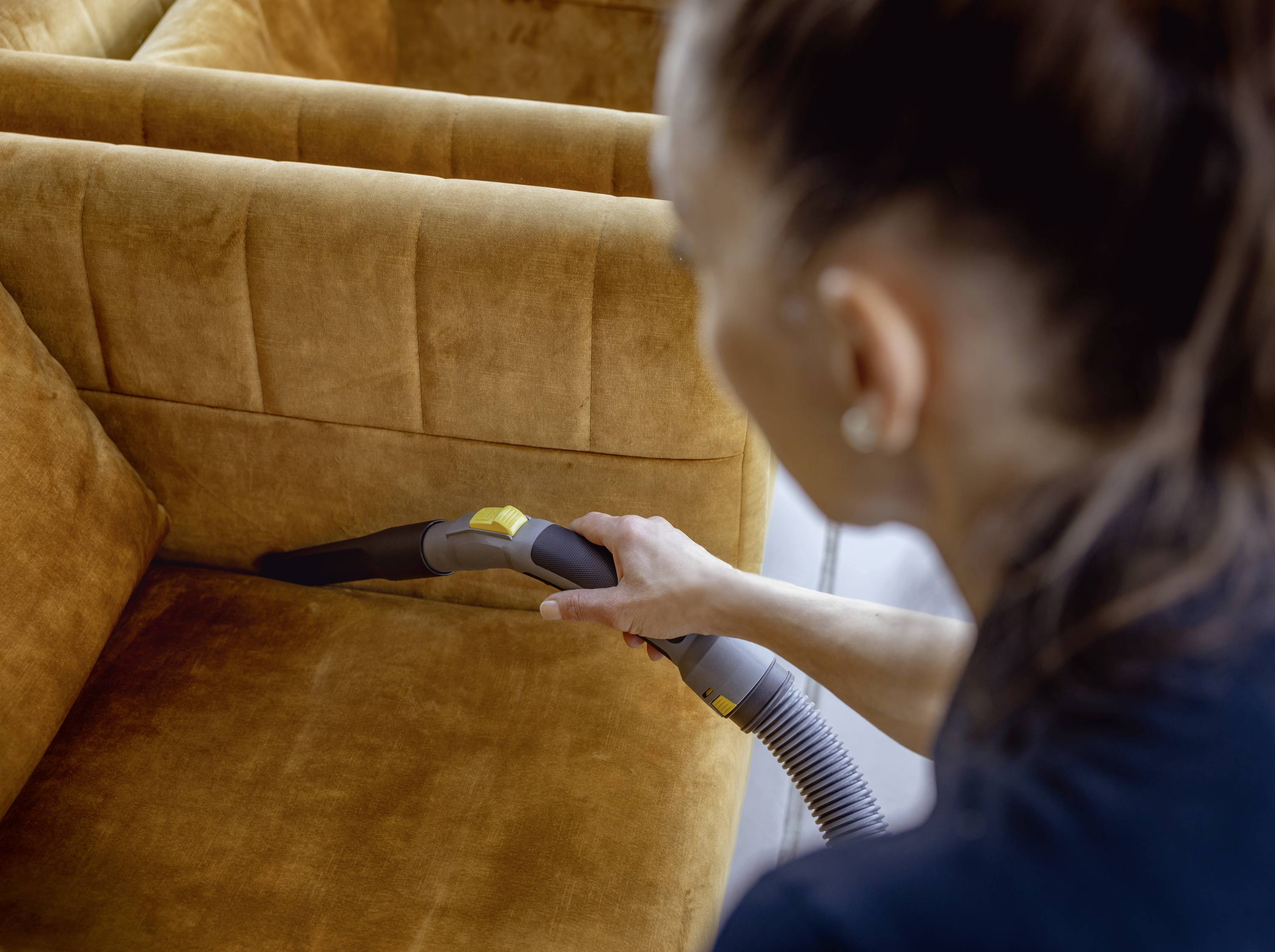 Eine Person saugt Sofapolster mit einem Staubsauger ab. Fokus auf die Reinigung der Ritzen im senfgelben Stoffsofa.