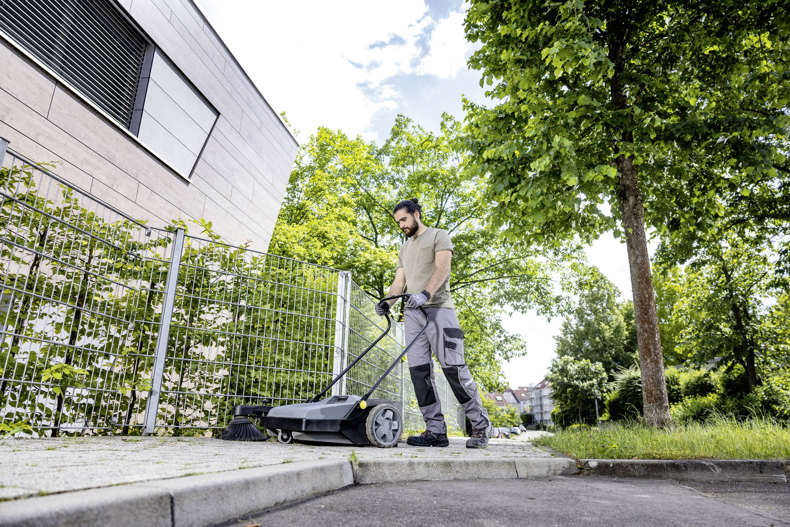 Ein Mann kehrt mit einer Kehrmaschine den Gehweg entlang eines modernen Gebäudes. Bäume säumen den Rand des Gehwegs.