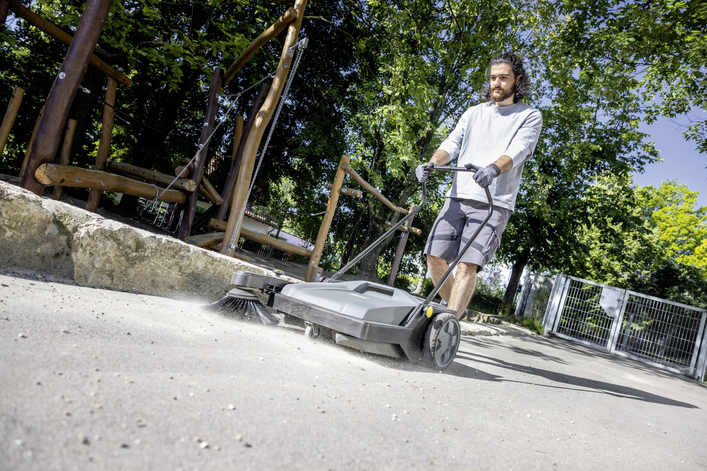 Ein Mann kehrt mit einer Kehrmaschine einen Gehweg im Freien. Im Hintergrund sind Bäume und ein Zaun zu sehen.