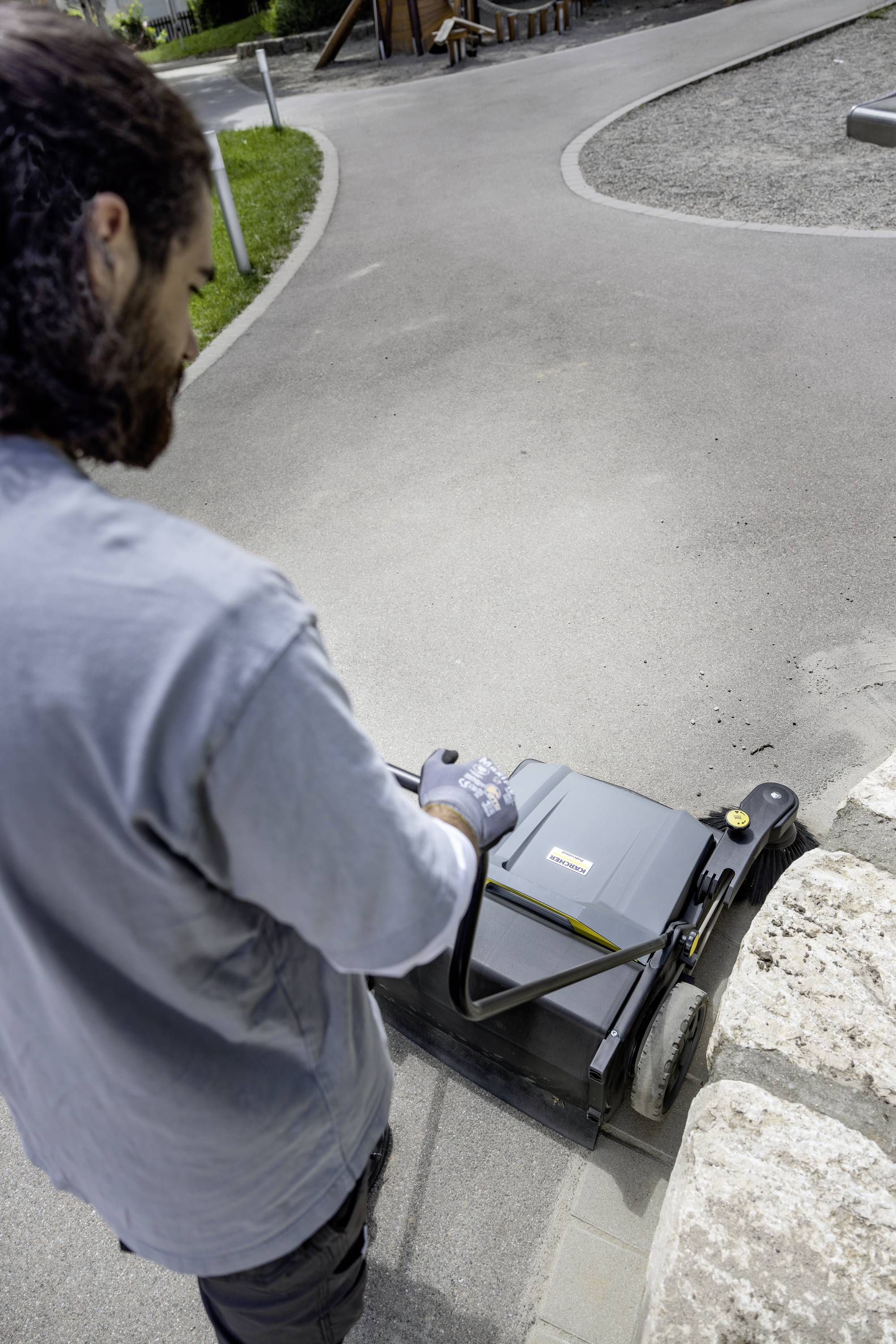 Eine Person bedient eine Bodenkehrmaschine auf einem Gehweg neben einer Mauer. Im Hintergrund ist ein Gartenbereich zu sehen.