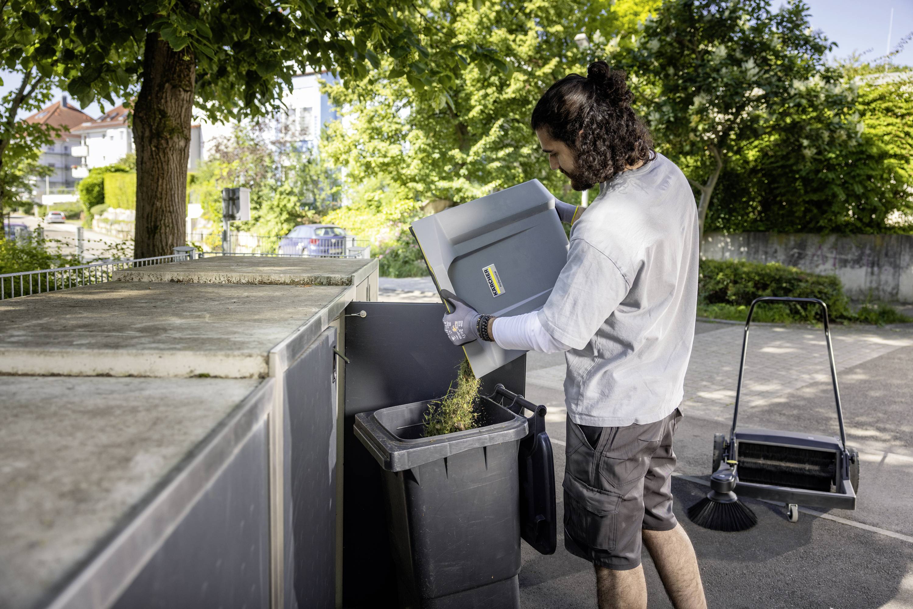 Eine Person entsorgt Pappe in einen großen Müllcontainer im Freien. Im Hintergrund sind Bäume und ein Gehweg zu sehen.
