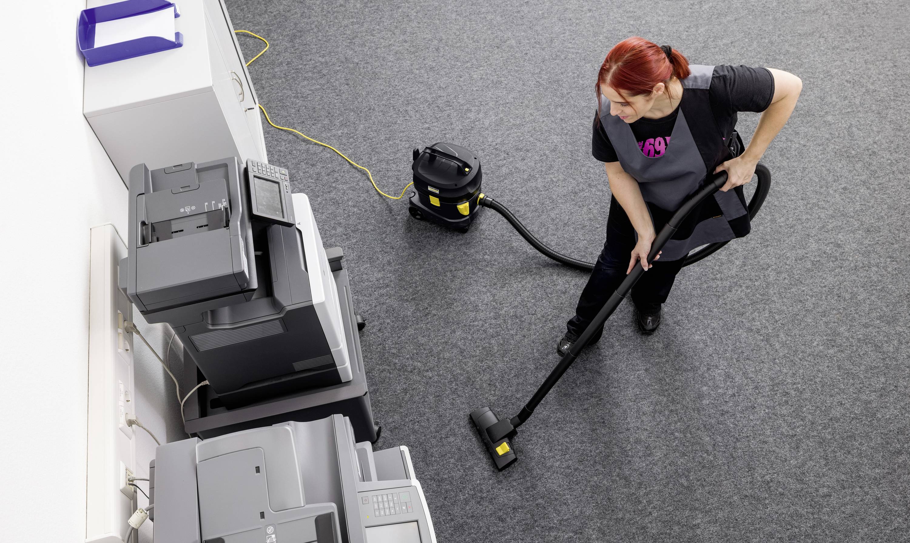 Eine Person mit einem Staubsauger reinigt einen Teppichboden in einem Büro mit mehreren Kopiergeräten an einer Wand.