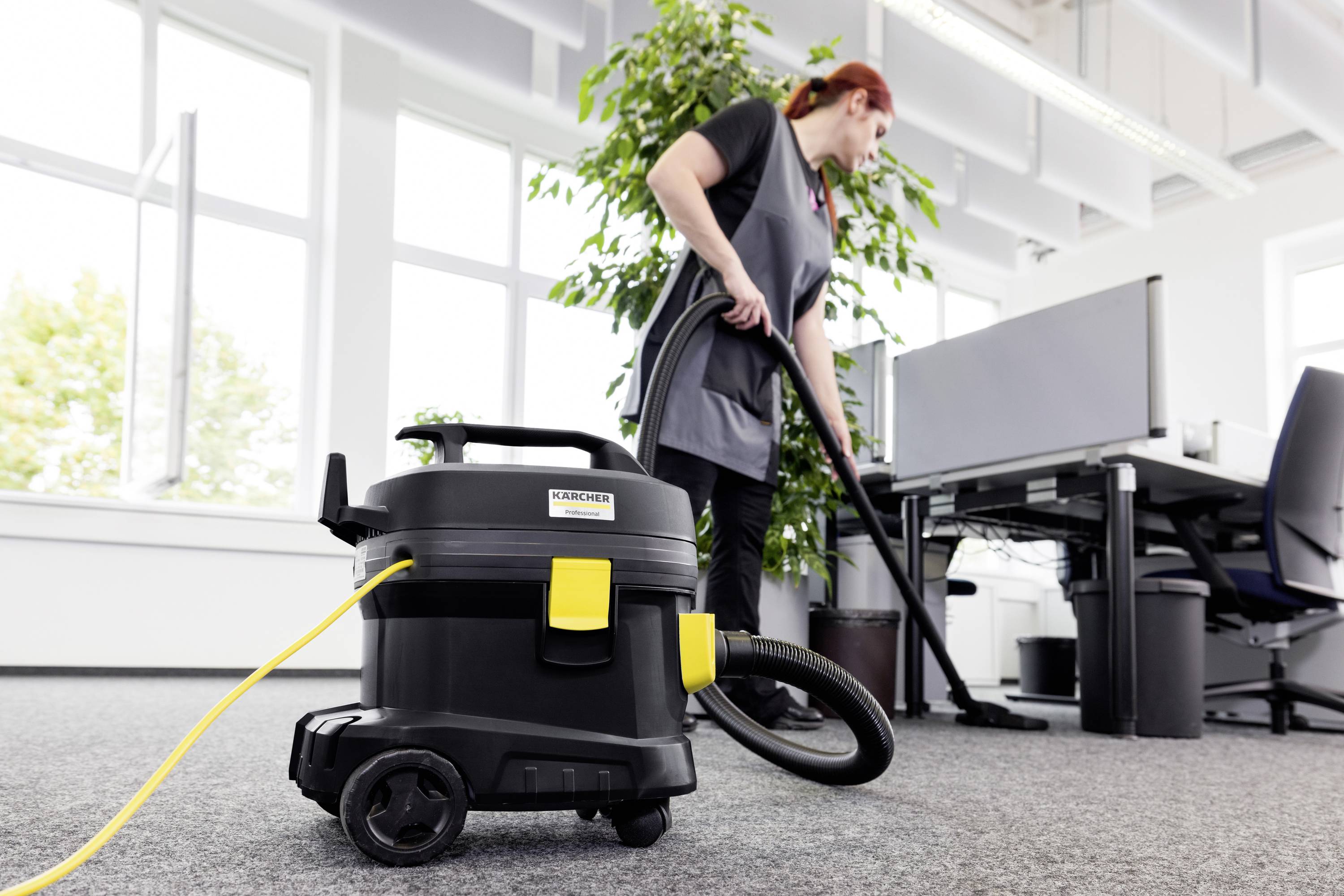 Eine Frau saugt ein Büro mit einem Industriestaubsauger. Im Hintergrund stehen Schreibtische und ein großer grüner Pflanztopf.