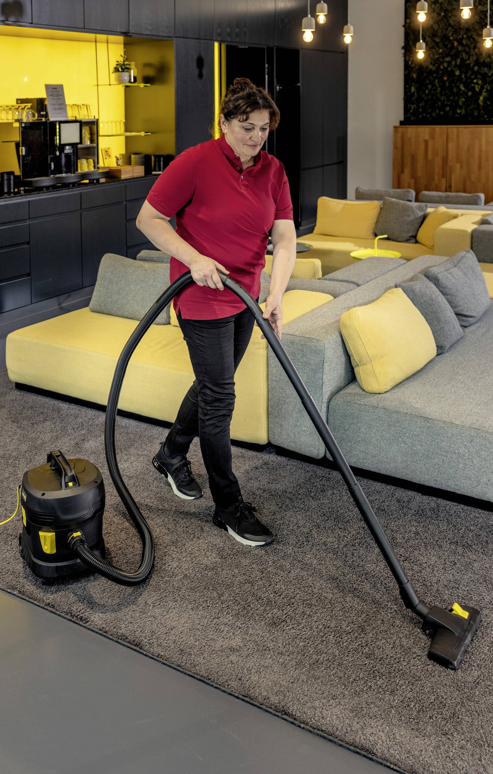 Eine Person in einem roten Shirt saugt einen mit Teppich ausgelegten Boden in einem modernen Wohnzimmer mit gelben und grauen Möbeln.