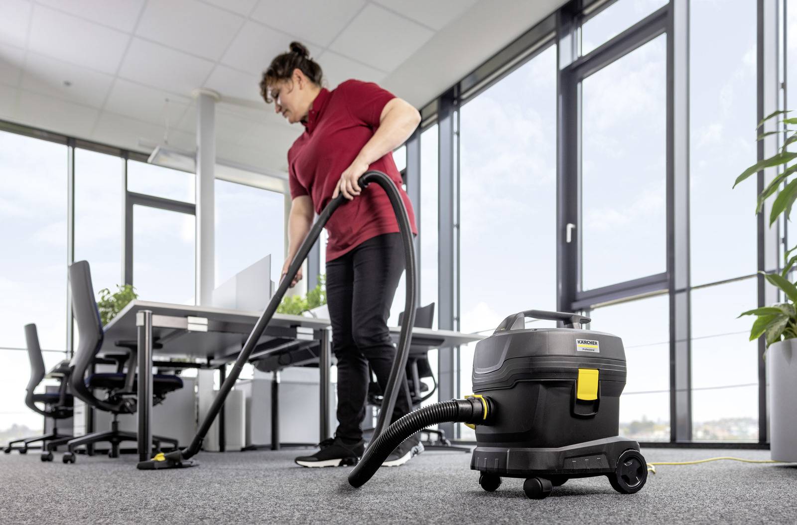 Eine Person in einem roten Hemd saugt ein Büro mit großen Fenstern mit einem Bodenstaubsauger auf einem Teppichboden.