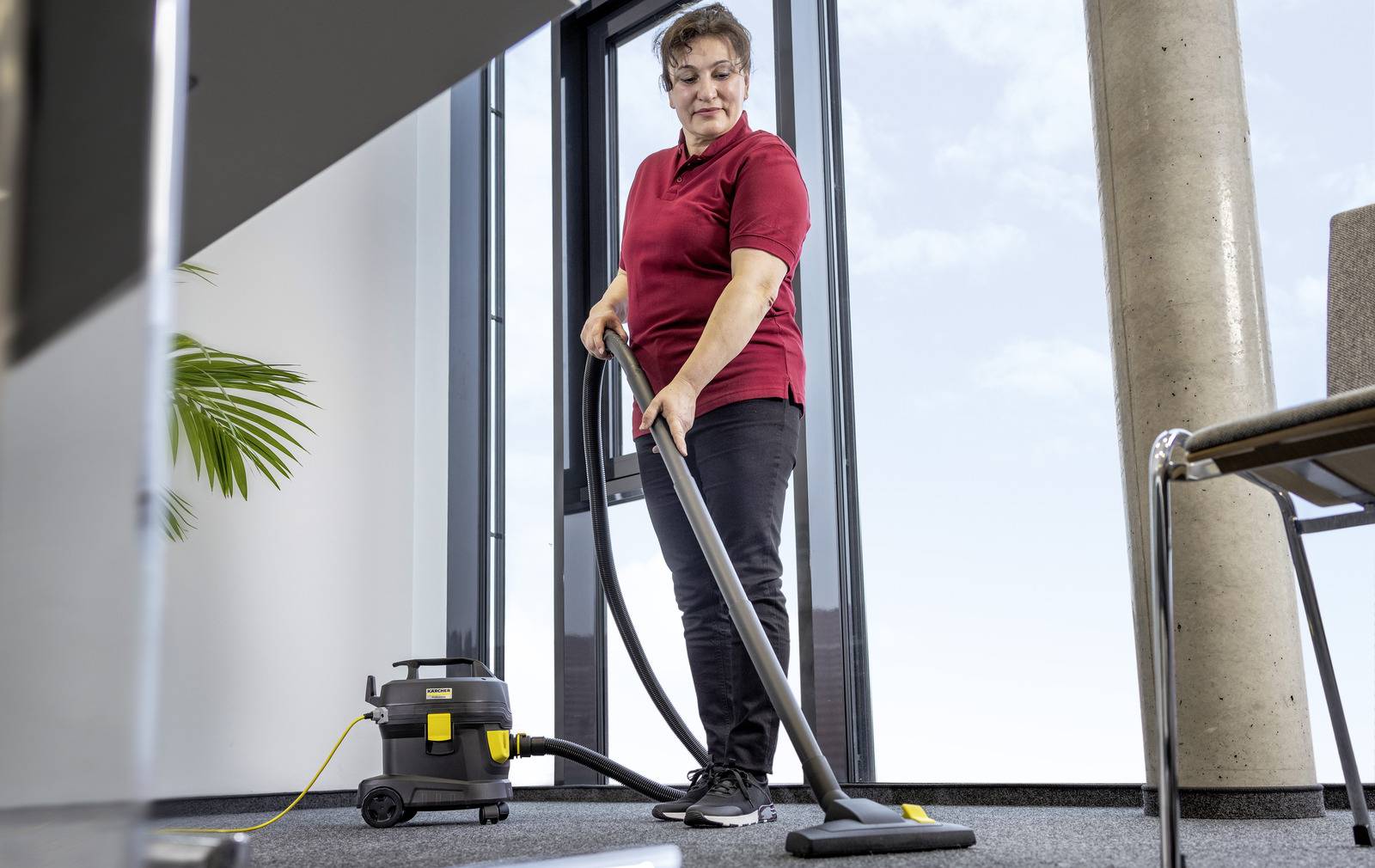 Eine Person in einem roten Hemd saugt einen Teppichboden in einem modernen Büroraum, wobei ein Fenster und eine Pflanze im Hintergrund sichtbar sind.
