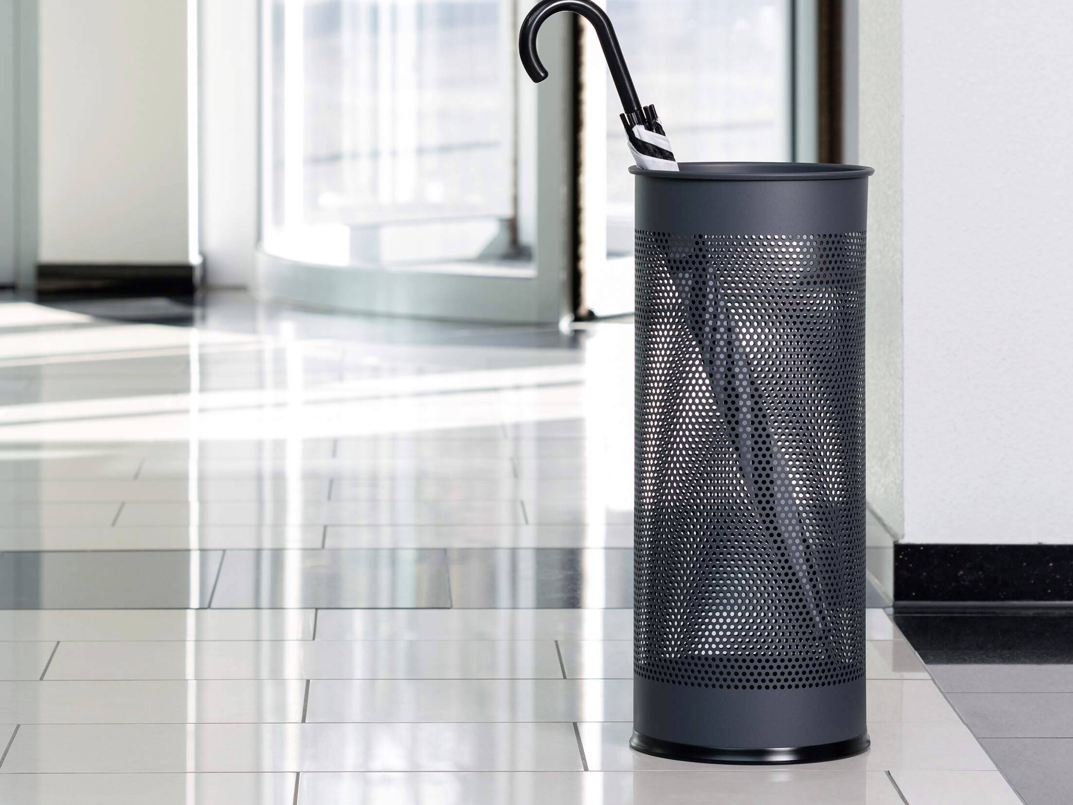 Ein schwarzer Schirmständer steht im Foyer auf glänzenden Fliesen, enthält einen zusammengefalteten Regenschirm.