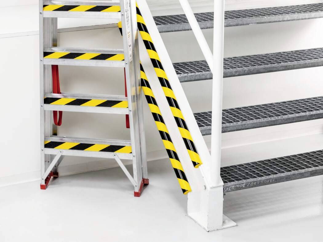 Metalltreppe mit schwarz-gelben Sicherheitsstreifen an Geländer und Stufen, roter Handlauf, weiße Wände, grauer Boden.